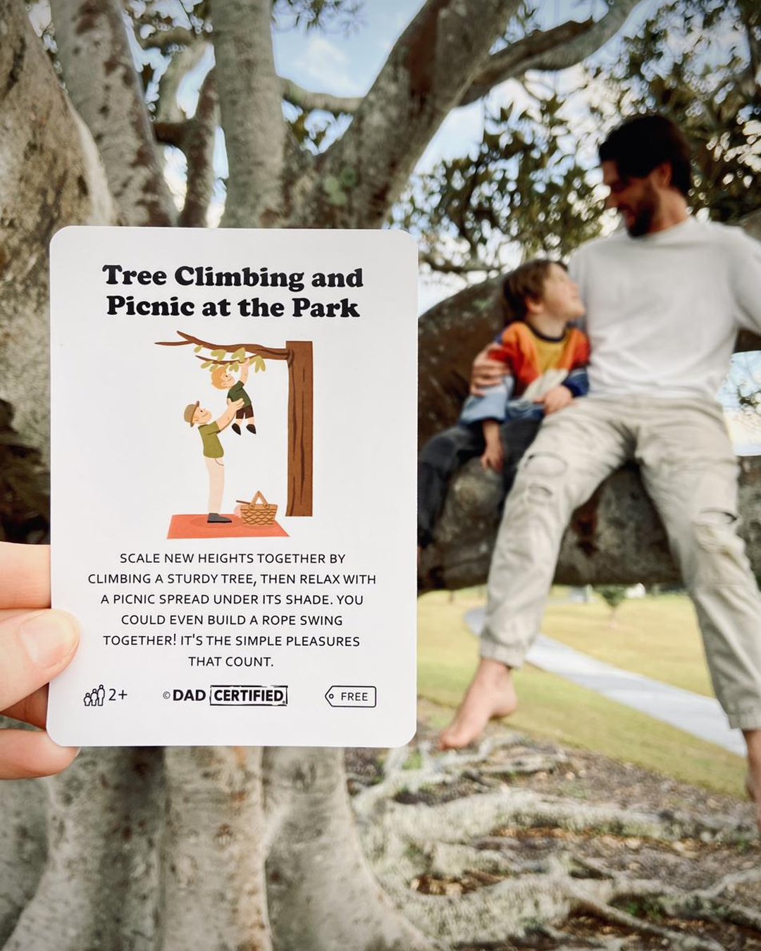 A dad holds the Tree Climbing Dadventure card while sitting in a tree with his son, living the exact activity on the card